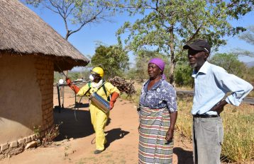 Spray campaign in Zimbabwe showing a rural home with a thatched roof on the left and a spray operator in yellow PPE and a woman with a purple head scarf and a man with a baseball cap on the right side and trees in the background and blue sky behind.