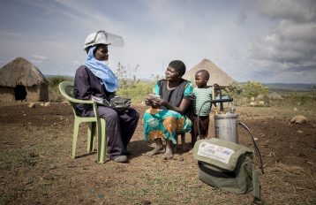 IRS sprayer sitting in a green chair wearing protective equipment and speaking with a mother and child outside of a rural home with a thatched roof