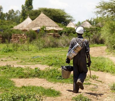 Man carrying indoor residual spraying equipment walking towards rural houses