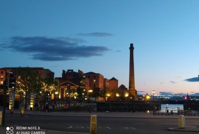 Photograph of the Albert Docks in Liverpool, UK at sunset, showing buildings to the left with the pump house tower with the road at the forefront and the sky deep blue tinged with red and a single cloud in the sky