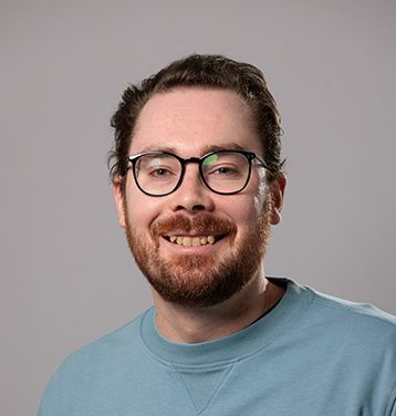 Image of a smiling, brown haired main with a red/brown beard and black rimmed glasses wearing a light blue sweatshirt. Background is grey.