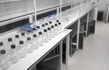 White lab bench diagonal from left to right across the image with two long rows of glass bottles with black lids in the forefront of the image and paper cups with netting in the back with a blue lab notebook