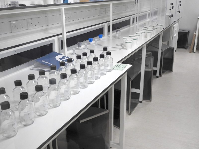 White lab bench diagonal from left to right across the image with two long rows of glass bottles with black lids in the forefront of the image and paper cups with netting in the back with a blue lab notebook