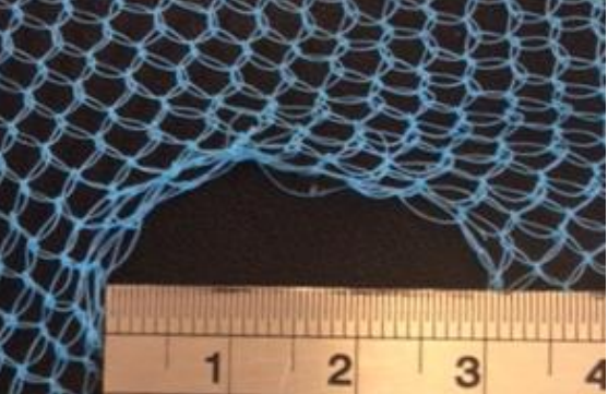 Image of blue netting against a black background with a hole in the bottom centre of the net fabric. A silver ruler at the bottom of the image is laid across the diameter of the hole, showing the hole is 3 cm wide.