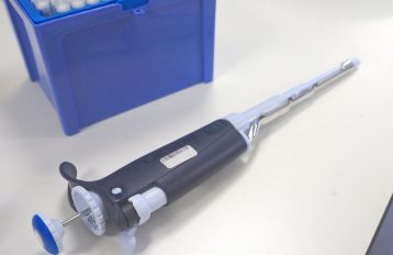 Open blue pipette tip box in upper left corner of image with dark blue and white pipette without tip lying on a white lab table