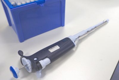 Open blue pipette tip box in upper left corner of image with dark blue and white pipette without tip lying on a white lab table