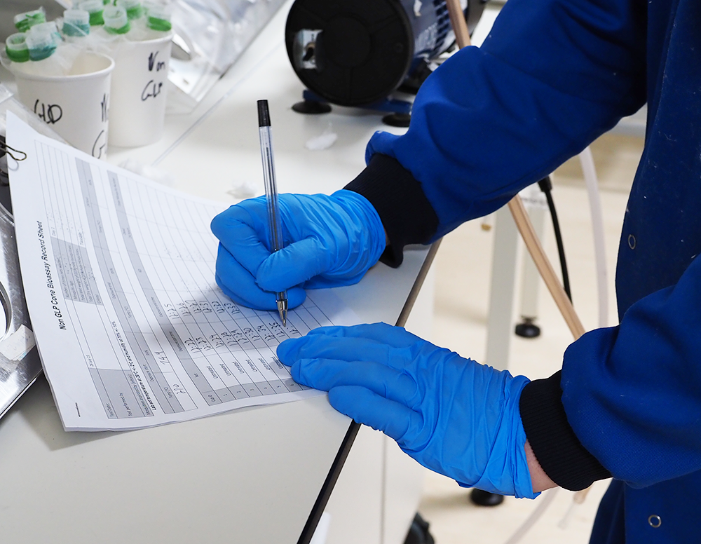 Picture of two hands in latex blue gloves of person wearing a royal blue lab coat. The left hand is holding a data collection sheet in place on a lab counter on the left of the image. The right hand is using a black pen to write data entries. Behind the data collection sheet is a white cup holding tubes with green rubber bands around the top of the tubes.
