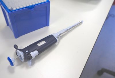 Bright blue open box of pipette tips in upper left corner with a dark blue and white pipette diagonally in front the box on a white lab bench counter.