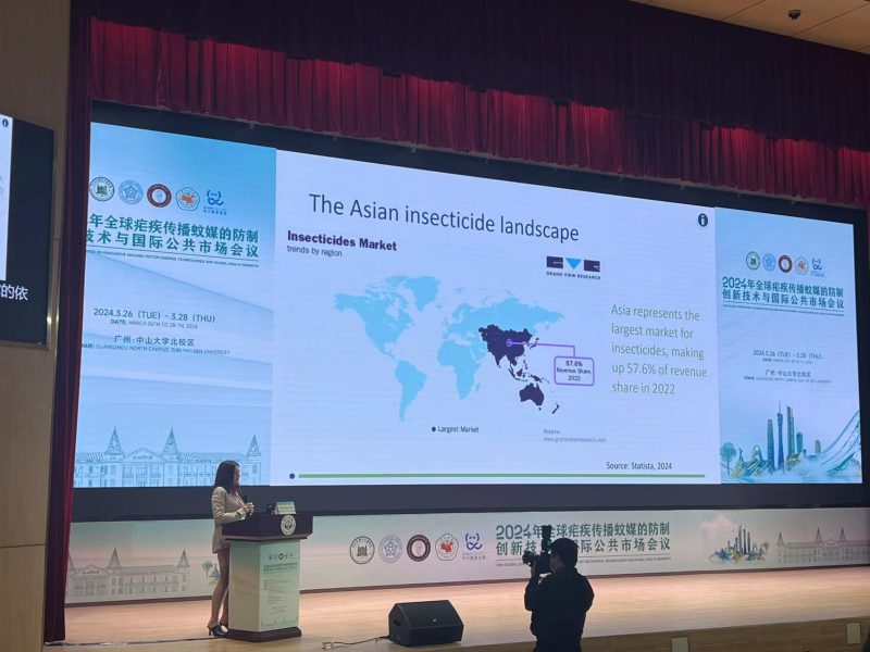 Large screen above a stage where a woman stands at the left of the screen at a podium. The screen displays a global map with the title 'The Asian insecticide landscape'.