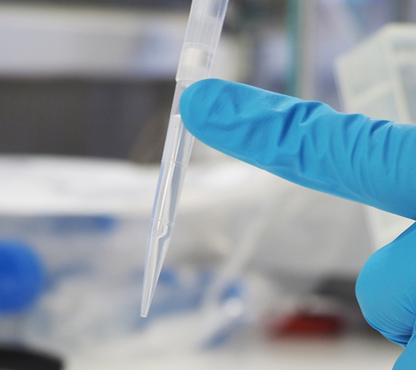 Pipette tip with clear liquid at centre of image, held in place by an index finger in a blue latex glove