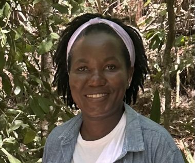 Picture of a woman smiling and wearing a white headband and a white t shirt with a blue collared overshirt. In the background is green foliage.